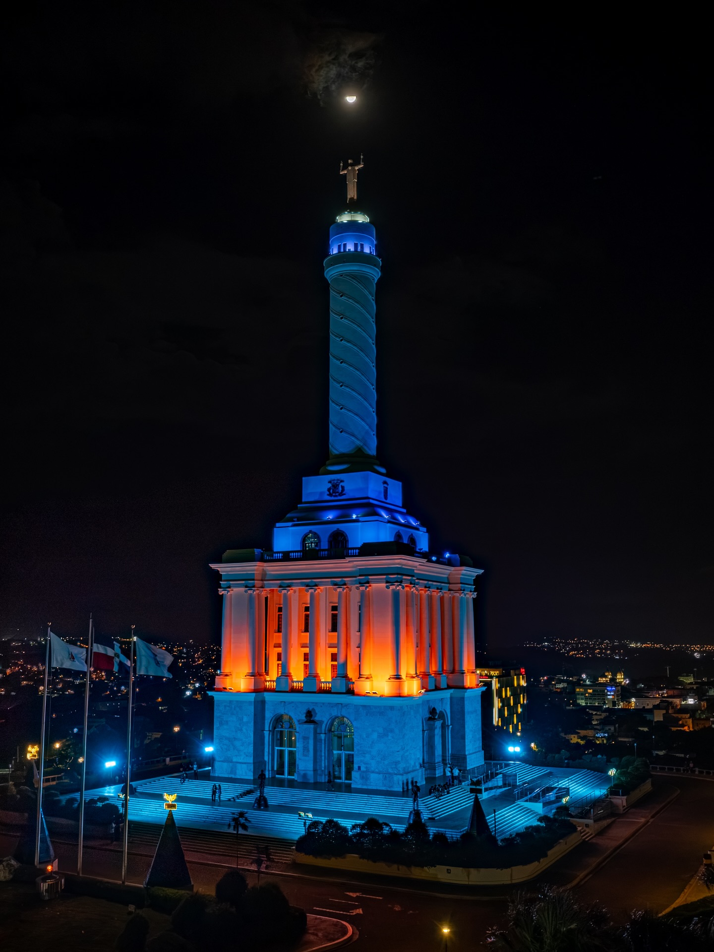 A monument illuminated in blue and orange beneath the moon at night.